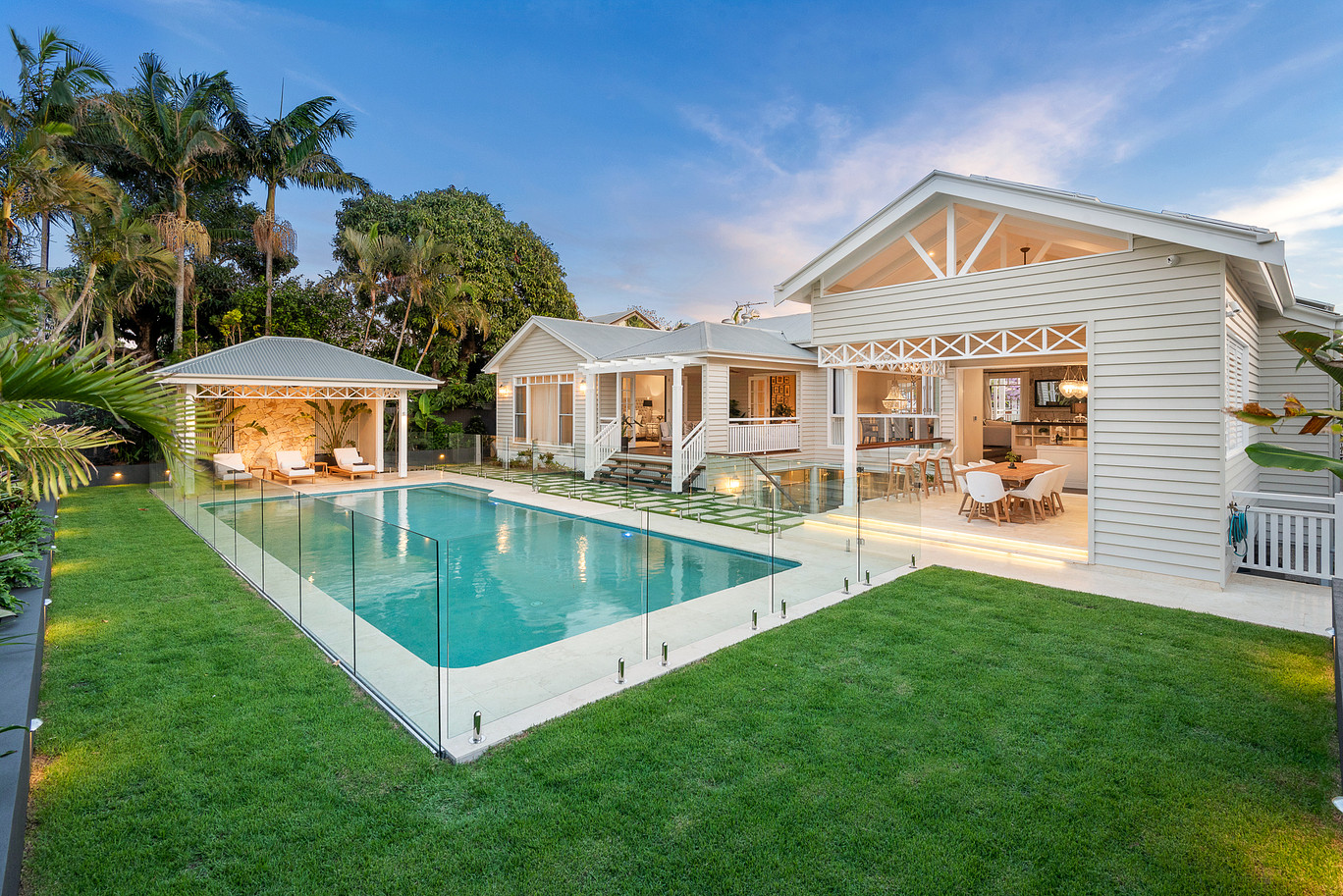 A modern Brisbane residence featuring a frameless glass pool fence with stainless steel spigots surrounding a lit swimming pool and manicured lawn.